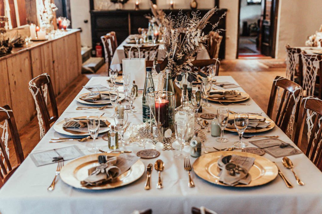 Tischdeko bei einer zweisprachigen Scheunenhochzeit im Bohostil im Nemsdorfer Hofgarten bei Nürnberg: goldene Platzteller, goldenes Besteck, Trockenblumen, Leinenservietten, dunkelrot, gold, weiß, beige